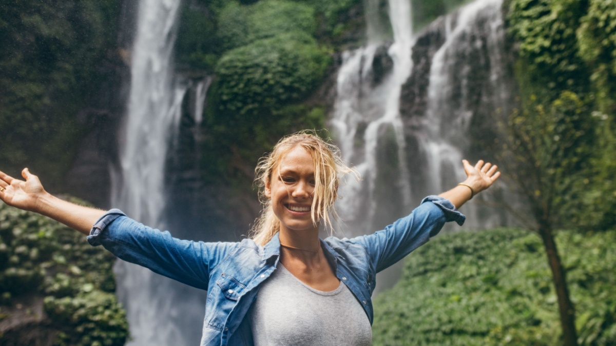 Portrait of young woman standing in front of a waterfall in forest with her hands outstretched. Caucasian female tourist with tropical waterfall in background.