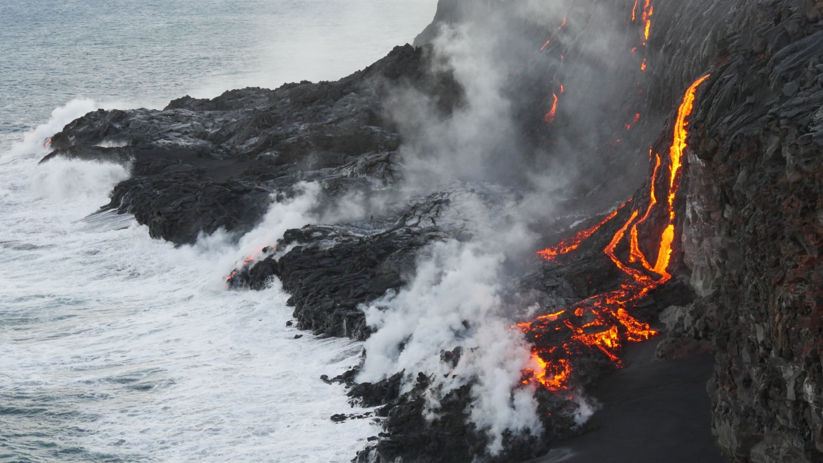 Molten lava flowing into the Pacific Ocean on Big Island of Hawaii, as ocean waves splash onto the rocks and flowing lava.