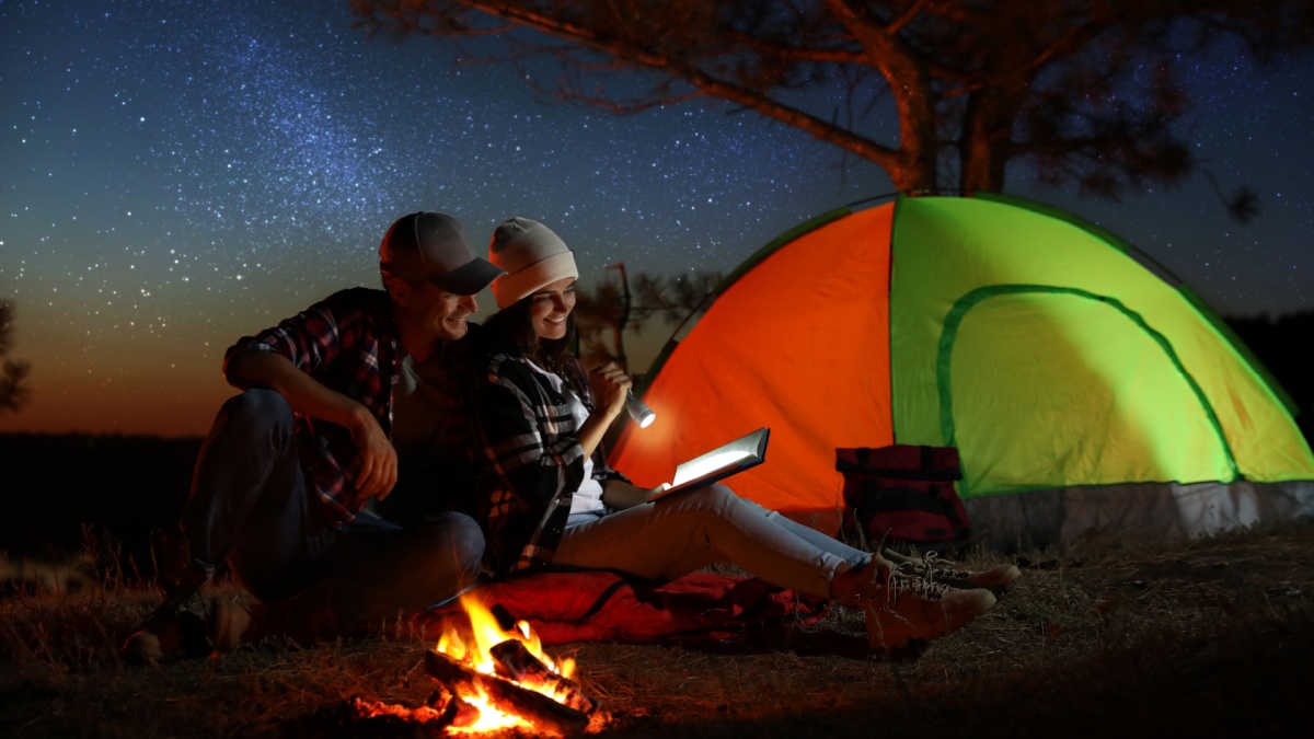 Couple with flashlight reading book near bonfire at night. Camping season.