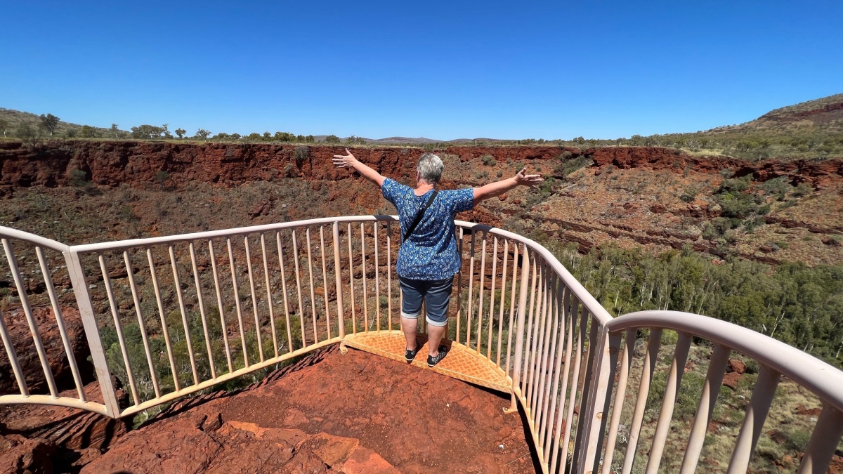 Mature aged female, with arms outstretched, viewing the spectacular landscape and gorges of Karijini National Park, Western Australia, from a red rock lookout and viewing platform.