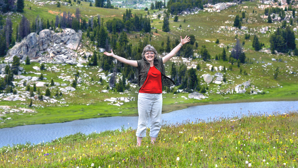 Woman spreads her arms wide to embrace the scenic beauty of Beartooth Pass in Wyoming. She has on a red shirt and khaki pants.