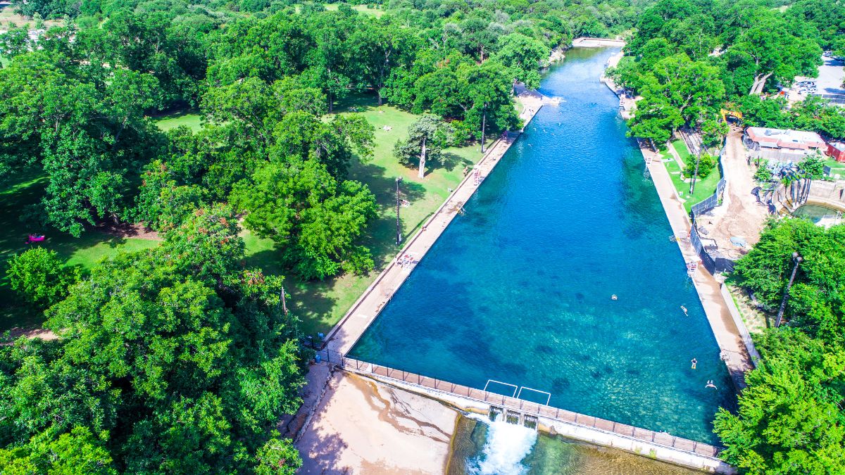 Looking down on Barton Springs natural cold spring swimming pool with dam and water rapids flowing off behind the one of a kind natural attraction of Austin Texas.