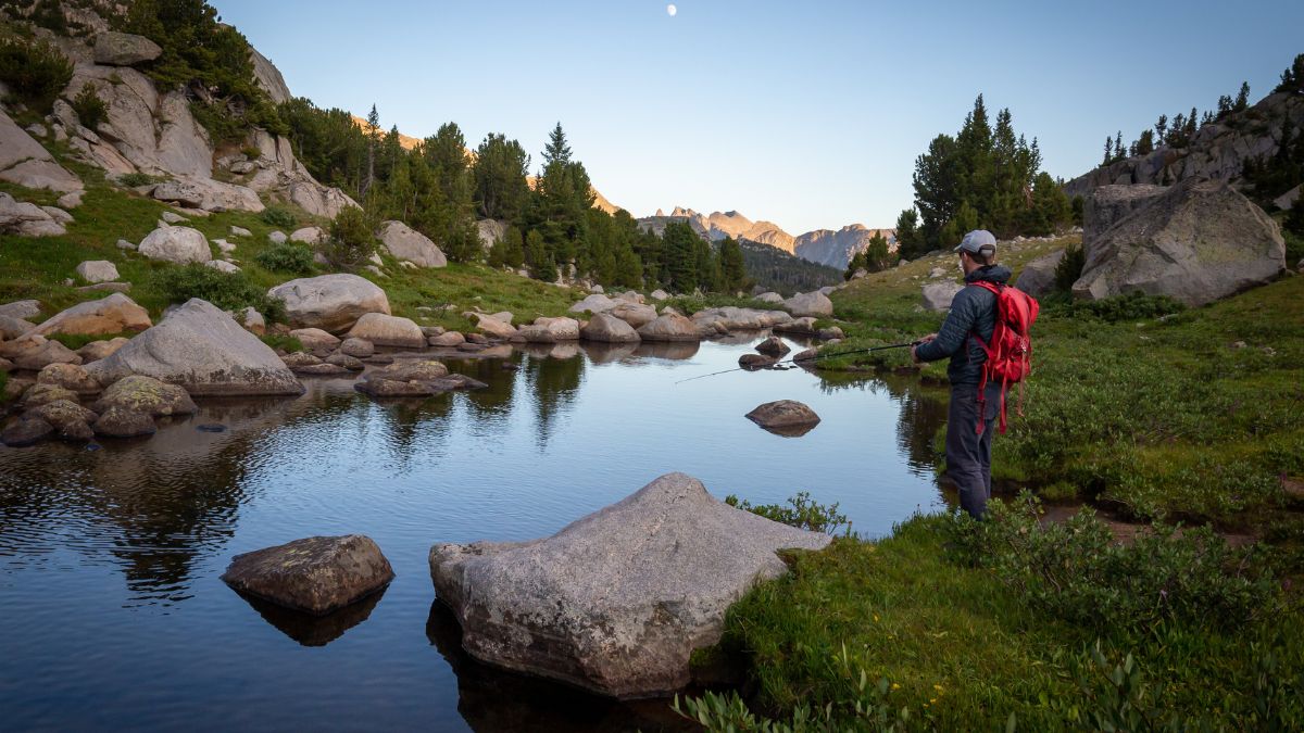 Fly fisherman in the Wind River Range in Wyoming fishing in a backcountry river stream during sunset with reflections on the water.