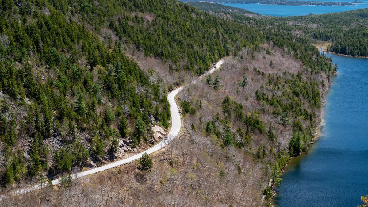 Winding Road Through Acadia National Park An aerial view of a winding paved road in Acadia National Park, Maine. The road snakes through a densely forested hillside, with a sparkling lake and ocean i.